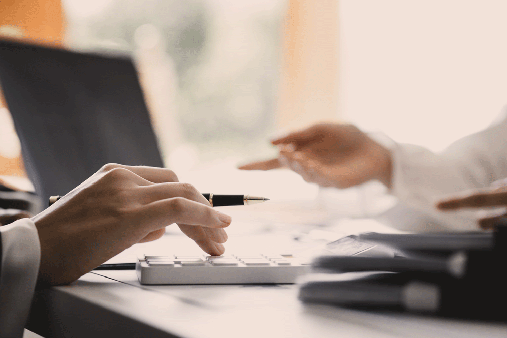 Two people sit at a desk reviewing documents; one uses a calculator or keyboard while the other gestures with an open hand. A laptop and a stack of folders are on the table, suggesting a collaborative business discussion.